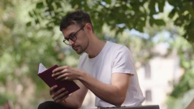 Genç adam parkta bankta oturur ve defterindeki notları okur. Eğitim, iş, planlar, fikirler, program. Beyaz tişörtlü adam..