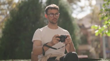 Genç bir adam, parkta bir bankta otururken çektiği fotoğrafları inceliyor. Bir fotoğraf daha çeker..