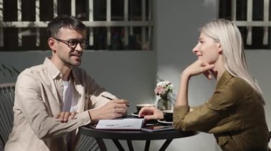 Bir adam ve bir kadın dışarıda bir kafede oturuyorlar, çalışıyorlar. Masanın üzerinde kahve ve notlarla dolu bir defter var. İş ortakları bir proje hakkında toplanıyorlar..