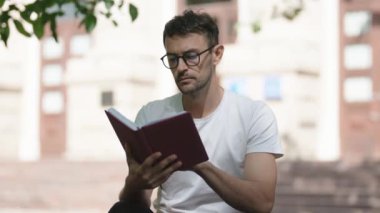 Parkta kitap okuyan bir adam. O, gözlük ve beyaz bir tişört giyiyor. Arkaplan bulanık ve bulanık.