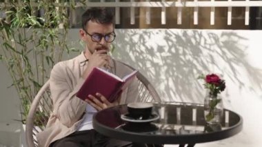 Deftere yazan ve açık kafede düşünen bir adam. O, gözlük ve bej bir gömlek giyiyor. Masanın üzerinde bir fincan kahve ve vazoda çiçekler var..