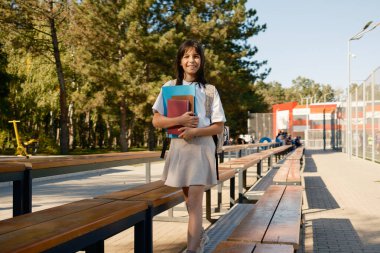 Okul parkında elinde kitaplarla neşeli bir genç öğrenci duruyor. Sahne huzurlu ve güneş ışığıyla yıkanmış akademik coşkunun özünü ve açık hava öğreniminin sükunetini yansıtıyor..
