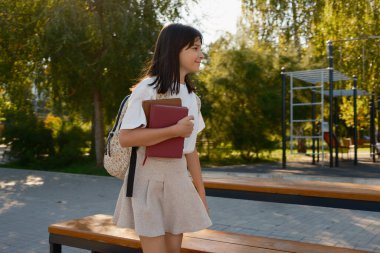 Genç bir öğrenci güneşli bir parkta yürürken kitap ve sırt çantası taşıyor. Neşeli ve yeşilliklerin arasında yeni bir öğrenme ve macera gününe hazır görünüyor..