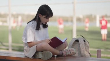Diğer çocuklar futbol oynarken, genç bir liseli kız bir bankta oturup okul spor sahasındaki derslerden sonra kitap okuyor. Eğitim, okuma, okul, öğrenme.