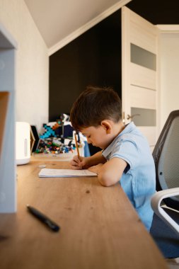Genç bir çocuk, rahat bir odadaki ahşap bir masada ev ödevini yapıyor. Sahne konsantrasyonu yakalar ve sıcak ve davetkar bir alanda çalışır..