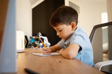 Genç bir çocuk, yumuşak ışıklandırmalı temiz bir masada oturan ev ödevine konsantre olur. Çevre sakin, odaklanma ve öğrenme deneyimini geliştiriyor. Eğitim ve öğrenim temaları için uygundur.