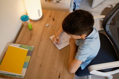 Genç bir çocuk, not defterleri ve bir lamba ile çevrili, masasında çizim yaparken konsantre olur. Sahne yaratıcılığı ve odağı aktarıyor, bir çocuğun öğrenme ortamını vurguluyor.