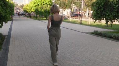 Genç bir kadın şehir kaldırımında kameradan uzaklaşıyor. O günlük giysiler giyiyor. Arka planda ağaçlar, çimenler ve arabalar var..