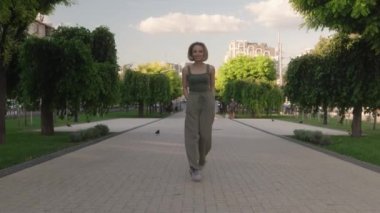 Sarı saçlı güzel bir genç kadın ağaçlarla kaplı bir şehir parkında kameraya doğru mutlu bir şekilde yürüyor. Duruyor, gülümsüyor ve yürümeye devam ediyor..