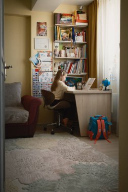 A young child is seated at a desk in a cozy home setting, studying. Bookshelves are organized behind the child, and natural light floods the room through the curtains, creating a serene atmosphere. clipart