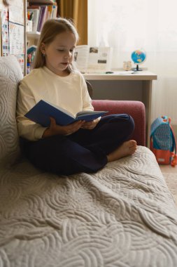 Bir kız kanepede otururken bir kitabın tadını çıkarıyor. Oda rahat, arka planda bir masa ve küre var, çalışma ve rahatlama vurgulanıyor..