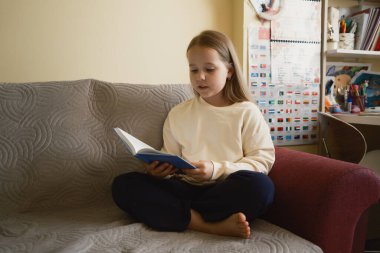 Genç bir kız kanepede rahatça oturup kitap okuyor, odaklandığını ve sakinliğini gösteriyor. Oda dekorasyonu sıcaklık ve yaşanabilir bir atmosfer ekler.