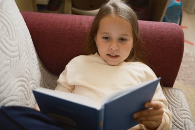 Genç bir kız evdeki rahat bir kanepede otururken dikkatle kitap okuyor, hem öğrenme hem de rahatlık sağlıyor..