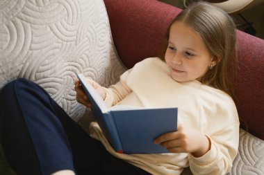 Genç bir kız rahat bir kanepede otururken kitap okumaktan zevk alır. Kurulumda rahatlama, eğitim ve evde boş vakitlerde okuma zevki vurgulanıyor..