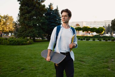 Kaykay ve not defteri taşıyan genç bir öğrenci güneşli bir parkta güvenle yürüyor..