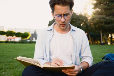Gözlüklü genç bir adam parkta çimlerin üzerinde otururken kitap okur. Sakin bir açık hava ortamı ve düşünceli ifadesi güneşli bir günde odak ve rahatlama sağlar..