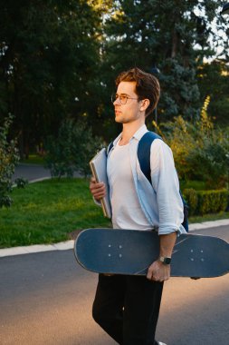 Elinde kaykay ve dizüstü bilgisayar tutan sıradan giyimli genç bir adam yeşil alanlarla çevrili bir parkta geziniyor. Gözlük takıyor, bir sırt çantası, ve açık havada eğlenirken düşüncelerde kaybolmuş gibi görünüyor..