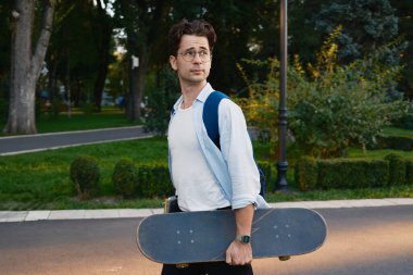 Gün batımında yeşil bir parkta elinde kaykay tutan genç bir adam, aktif ve rahat bir yaşam tarzını yansıtıyor..