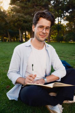 Genç bir adam çimlerin üzerinde bağdaş kurup defterine bir şeyler yazıyor. Sakin açık hava koşulları ve odaklanmış tavırları bir konsantrasyon ve sakinlik ortamı yaratıyor..