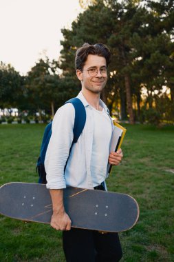 Açık hava manzarasının tadını çıkarırken elinde kaykay ve sırt çantası tutan kendine güvenen genç bir adam. Gençlik, hobiler, yaşam tarzı ve özgürlük temaları için mükemmel.