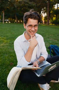 Neşeli genç adam dışarıda dizüstü bilgisayar ve kitap kullanarak güneşli bir günün tadını çıkarıyor. Doğanın ortasında üretken ve pozitif bir öğrenme ortamı yakalıyor..