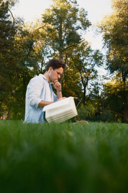 Parkta çimlerin üzerinde oturan bir adam, elinde bir kitapla çalışıyor, yeşilliklerle çevrili, güneşli havanın tadını çıkarıyor. Açık havada huzurlu ve üretken bir an..