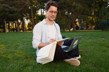 Bir erkek öğrenci parkta dizüstü bilgisayar kullanırken deftere notlar yazıyor. Sahne, ağaçlar ve yeşillikler arasında bir açık hava ortamında rahatlama, öğrenme ve verimliliği tasvir ediyor..