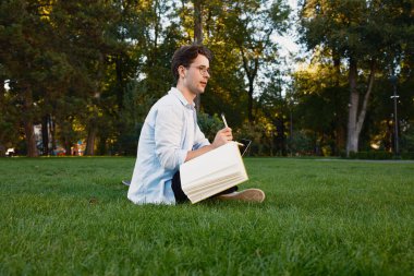 Sıradan giyinmiş genç bir adam, parkta bir defterle çimlerin üzerinde oturuyor. Yeşillik ve güneş ışığıyla çevrili, yazarken ya da çizerken düşünceli ve meşgul görünüyor..