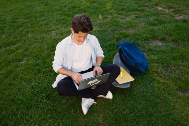 Doğa tarafından çevrelenmiş, dışarıda dizüstü bilgisayarla ders çalışan sıradan bir öğrenci. Rahatlamış atmosfer, kaykay, defter ve sırt çantasıyla vurgulanarak çimenlerin üzerinde rahat bir çalışma alanı yaratıyor.