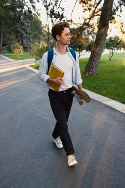 Sıradan giyinmiş genç bir adam, elinde kaykay ve defterlerle parka benzeyen bir kampüs ortamında yürüyor ve hoş akşam ortamının tadını çıkarıyor..