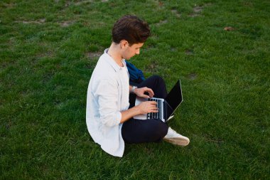 Genç adam dışarıda dizüstü bilgisayarıyla çalışıyor. Sıradan bir park ortamında yeşil çimlerin üzerinde oturuyor..