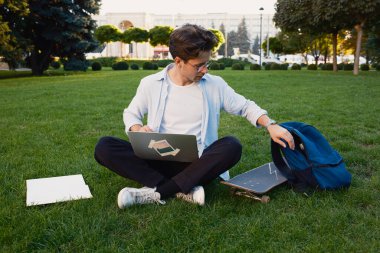 Bir öğrenci parkta çimlerin üzerinde dizüstü bilgisayar, kaykay ve sırt çantasıyla yan yana oturur. Rahatlamayı ve öğrenmeyi önerir..