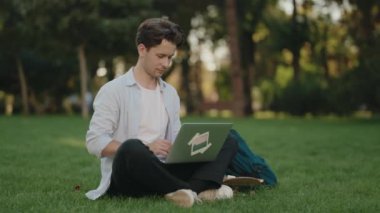 Genç bir adam parkta çimlerde oturur ve dizüstü bilgisayarıyla çalışır. Uzaktan çalışırken odaklanmış ve üretken görünüyor..