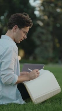 Genç bir adam parkta çimlerin üzerinde oturur ve büyük bir deftere yazar. Yanında gözlük ve laptop var..