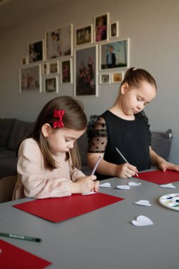 İki genç kız, arka planda çerçeveli fotoğraflarla, yaratıcılığı ve işbirliğini vurgulayan, kapalı bir alanda kırmızı kağıt kalplerle sanat ve zanaat yaratmaya odaklanıyor..