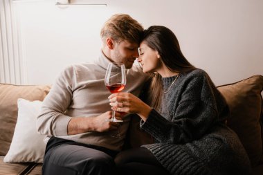Sevgi dolu bir çift evde samimi bir anı paylaşıyor, ellerinde kırmızı şarap kadehleri, sıcak ve rahat bir atmosfer yaratıyorlar. Mükemmel bir sevgi tasviri, rahatlama ve içten bir bağ..