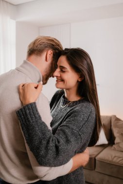 Rahat bir yaşam alanında bir çiftin kucaklaşmasını içeren samimi bir an. Sahne sıcaklığı, sevgiyi ve sevgiyi yansıtıyor, arka planda parlak, modern bir iç mekan var..