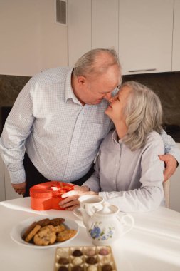 An elderly couple enjoying a heartfelt moment together, exchanging gifts and warm gestures, seated in a bright kitchen with treats and tea on the table, symbolizing love, care, and togetherness. clipart