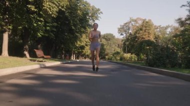 Genç bir kadın güneşli bir sabahta güzel yeşil bir parkta koşuya çıkar. Spor kıyafeti giyiyor ve hafif bir koşuda koşuyor. Temiz havanın tadını çıkarıyor..
