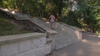 Genç bir kadın spor programının bir parçası olarak parkta uzun bir merdivenden yukarı koşuyor..