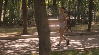 Genç bir kadın huzurlu bir parkta yürüyor. Spor kıyafetleri giyiyor. Egzersiz yapmaya ya da serinlemeye ara verdiğini gösteriyor..