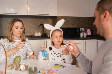 A family engaged in colorful Easter egg paintings at a modern kitchen table. clipart