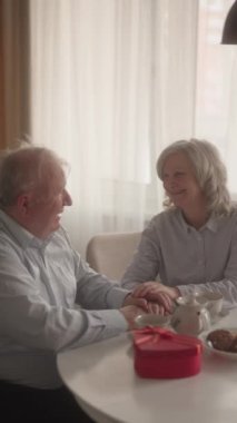 Yaşlı bir çift el ele tutuşuyor, gülümsüyor ve masada kahve içiyor..