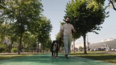 Bir adam güneşli bir günde Bernese Dağ Köpeğini ağaçlarla kaplı bir şehir parkında gezdirir.