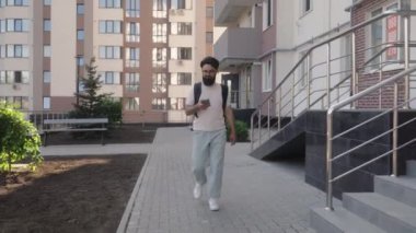 Sakallı ve gözlüklü genç bir adam dışarıda yürüyor ve akıllı telefon kullanıyor. Günlük giysiler ve sırt çantası giyiyor. Kentsel ortam ve doğal ışık modern ve anlaşılabilir bir sahne yaratır..