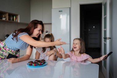 Bir anne esprili bir şekilde kızlarını mutfak masasına götürmeye çalışır. Etrafı keklerle çevrilidir. Eğlenceli ve hayat dolu bir aile anı yaratır..