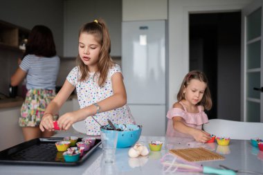 İki genç kız modern bir mutfakta renkli kekler pişirmekten, aile birlikteliği ve eğlencesini sergilemekten hoşlanıyorlar. Yaratıcı bir aşçılık faaliyeti içindeler..