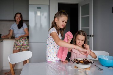 İki genç kız kardeş mutfakta birlikte yemek yapmaktan zevk alırken anneleri arka planda mutlu bir şekilde gülümsüyor. Sahne, sıcak bir ev ortamında aile bağlarını, takım çalışmalarını ve eğlenceyi yakalıyor..