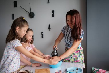 Bir anne ve iki kızı, çağdaş bir mutfakta eğlenceli bir yemek pişirme seansının keyfini çıkarıyorlar, neşe ve aile bağlarını paylaşıyorlar. Duvardaki saat modern bir dokunuş katıyor..