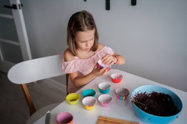 Genç bir kız masasında oturmuş, odak ve yaratıcılıkla renkli kek kapları düzenliyor. Bir kase çikolata hamuru pişirmeye hazır, eğlenceli bir ev yemeği anı yakalıyor..